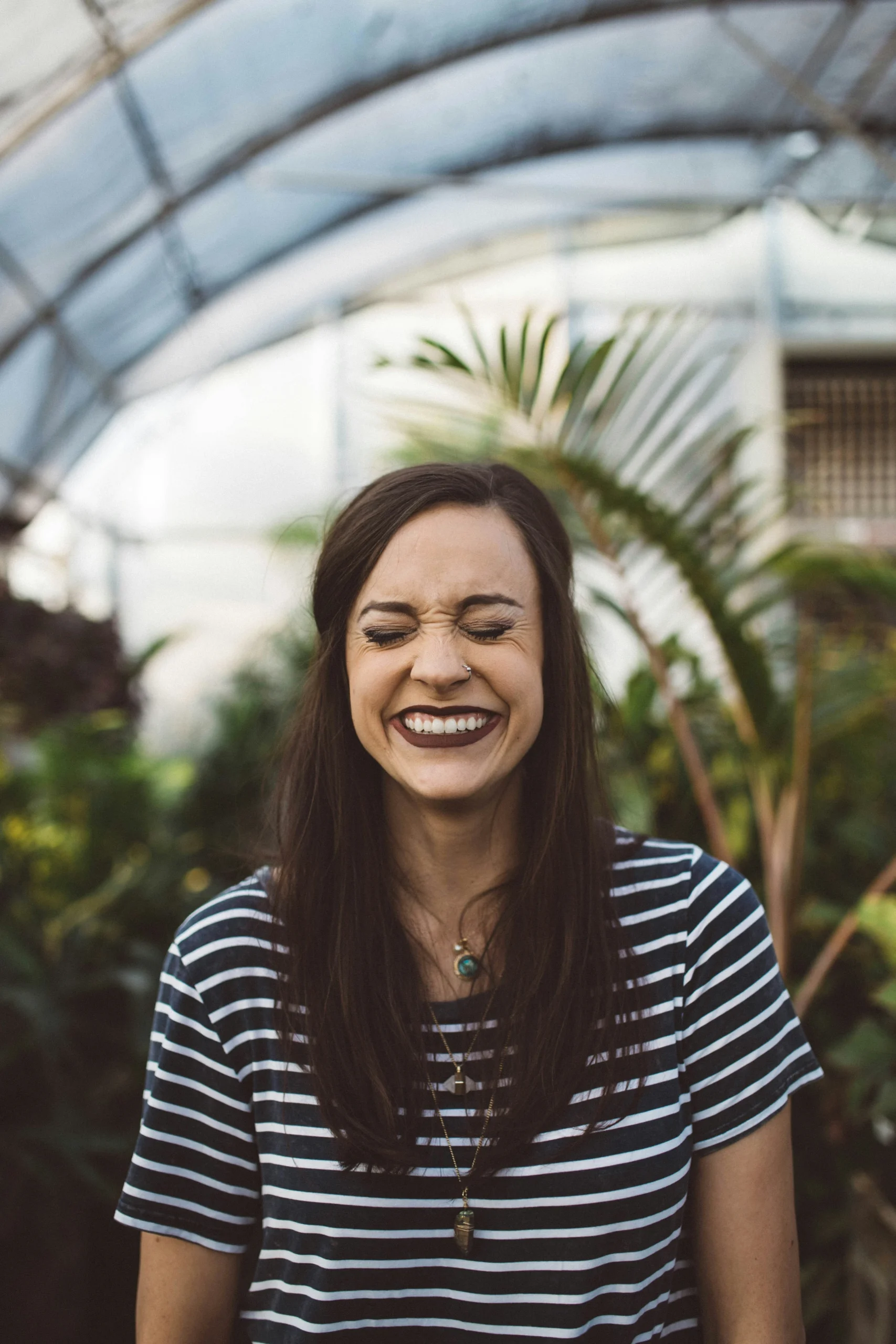 A woman mid laugh, wearing a horizontally striped shirt. In the background are plants.