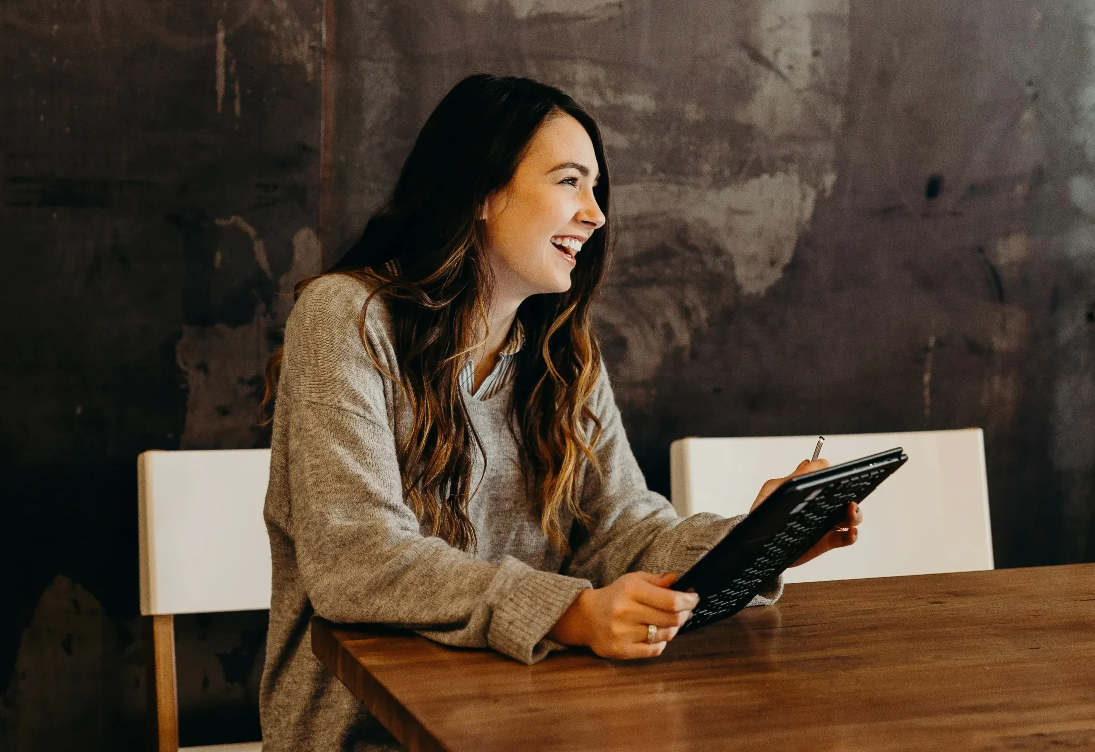 Smiling woman sitting at a wooden table holding a tablet, against a dark textured wall, exuding a cheerful and professional vibe.